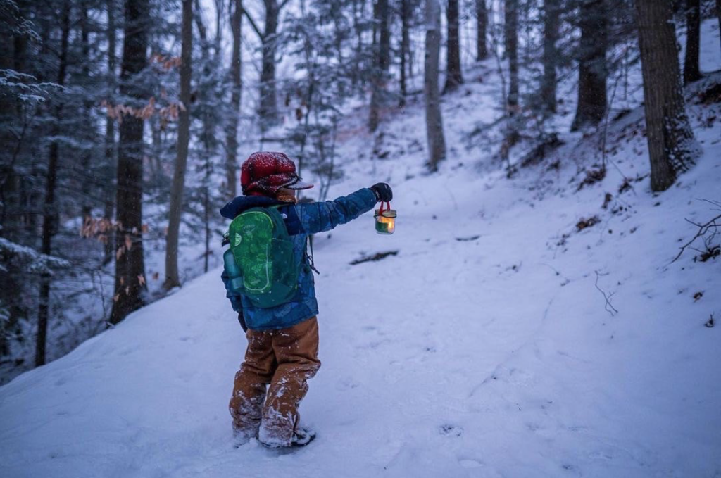 little boy with red hat, blue coat and green backpack hold lantern up while facing snow covered hill