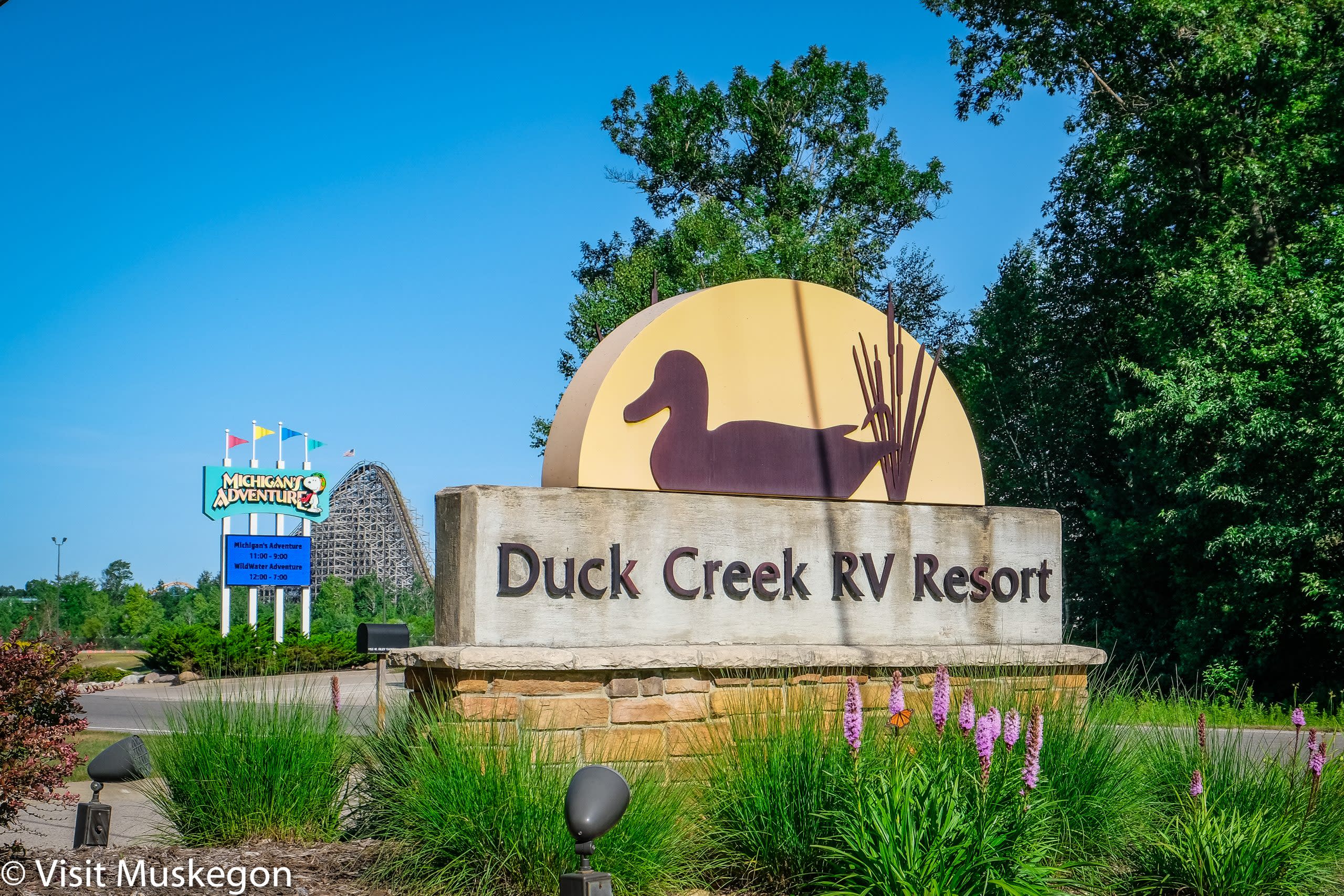 sign for duck creek RV resort sits among flowers, grass and trees. sign for michigans adventure and a rollercoaster are in background