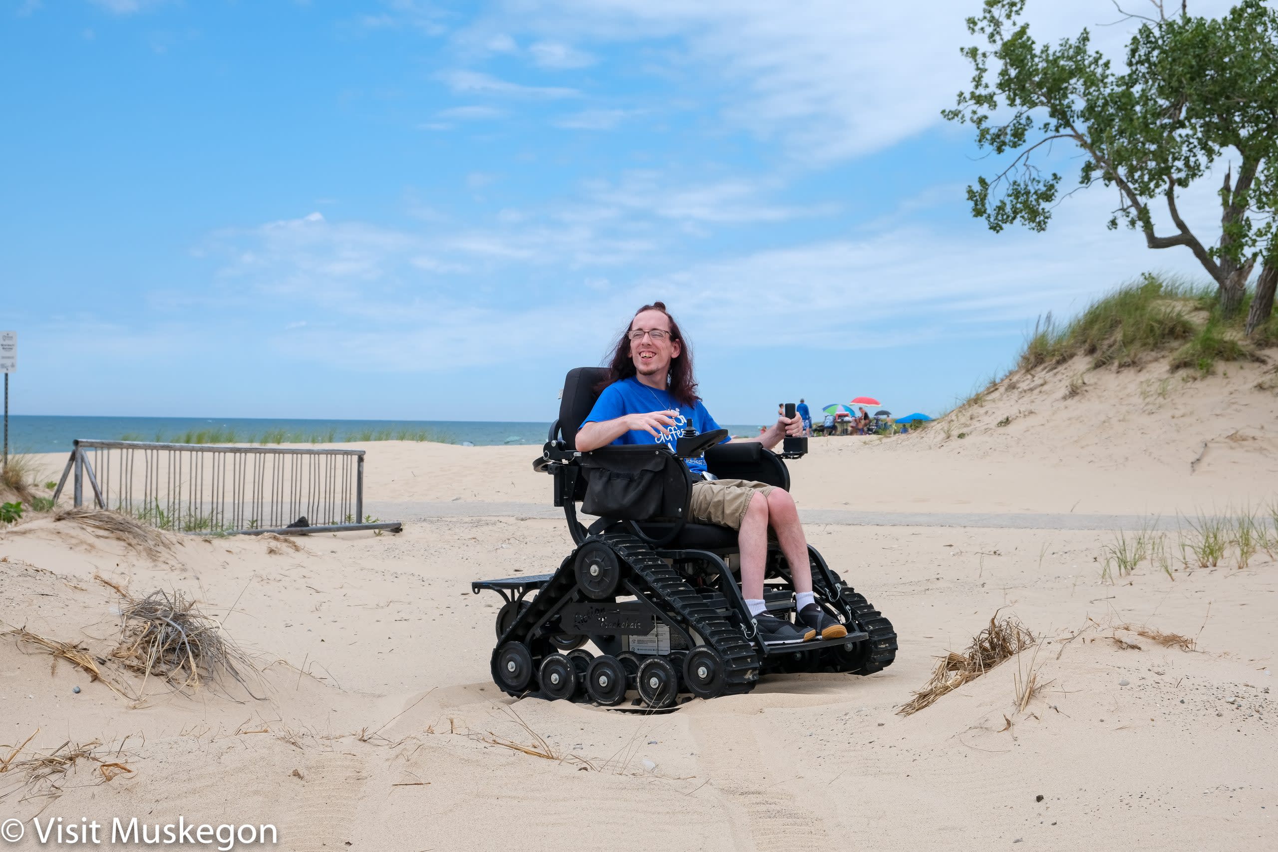 smiling young man in TrackChair on the beach. Lake Michigan is in the background and a small dune with a tree are to his right. 