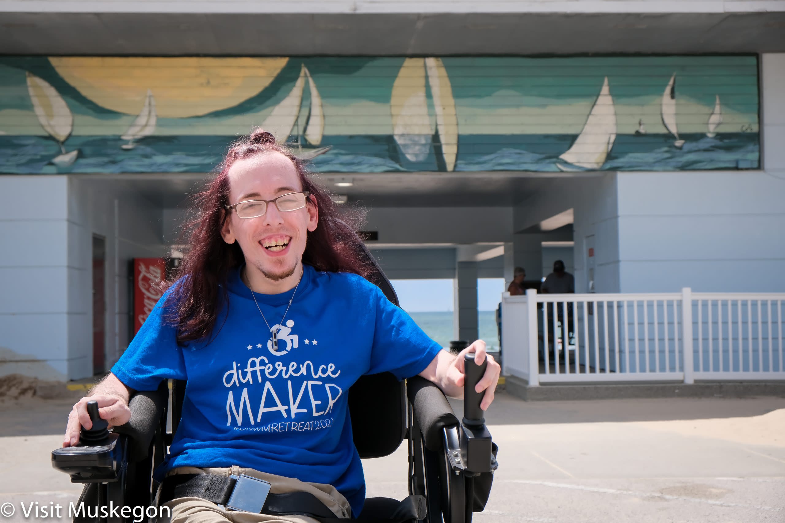 smiling young man is seated in a TrackChair with beach bathhouse in he background. His blue tee shirt says "difference maker. A mural of sailboats is above his head