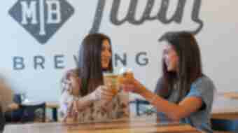 Two women cheers their beers and smile inside the tasting room of Mirror Twin Brewing.