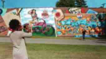 A woman photographs her friend posing for a photo in front of a series of huge, vibrant murals.