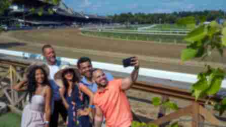 A group of people smiling and posing for a picture outdoors at the Saratoga Race Track, surrounded by trees and green grass under a clear sky.