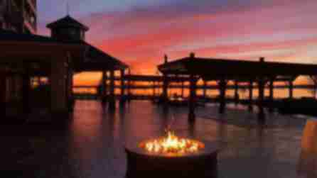 A view of the St. Lawrence River at sunset with orange, purple, and red skies from a fire pit with orange flame