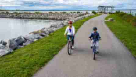 A woman and a child ride bicycles along the Empire Trail at its terminus at Buffalo Harbor State Park beside a calm lake, with rocky shores and a cloudy sky in the background.