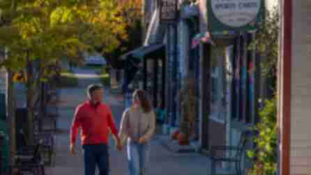 A couple walks hand-in-hand down a quaint, sunlit street lined with shops in autumn. Leaves are turning, pumpkins decorate storefronts, and they seem content.