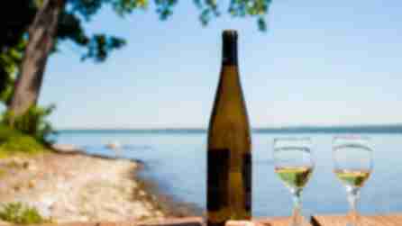 A bottle of wine sits next to two glasses of white wine on a wooden table in front of a view of Seneca lake