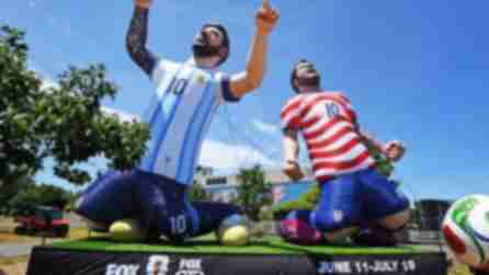 Two large inflatable soccer players kneeling, one in an Argentina jersey, the other in a USA jersey, with a giant ball nearby. Clear blue sky backdrop.