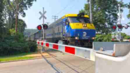 The blue and yellow front of the MTA LIRR train with the railroad crossing bar down on each side with green trees on either side of the train with the flashing red railroad crossing lights
