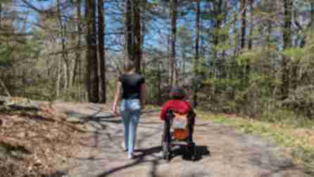 A person wearing jeans and a black shirt walks beside a wheelchair user wearing a red shirt on a forest path at Mohonk Mountain House.