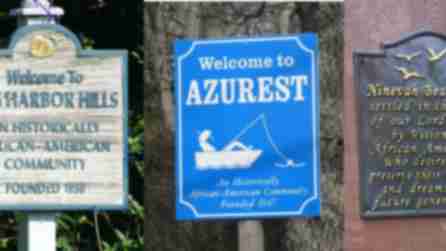 Three signs welcome visitors to historical African-American communities. "Sag Harbor Hills" is on a rustic wooden sign, "Azurest" on a bright blue sign with a boat graphic, and "Ninevah Beach" on a dark plaque with gold text and birds.