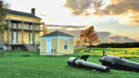 Two cannons resting on the field at Sackets Harbor Historic Site with a house and out-house in the background