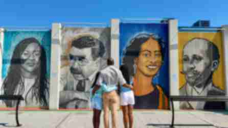 A family of three embraces while admiring the Freedom Wall in Buffalo, a collection of large, colorful murals featuring historical figures in a public outdoor space, evoking a sense of reflection.
