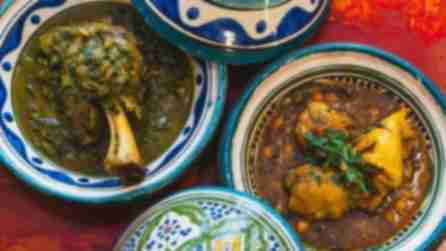 Overhead view of colorful Moroccan tagine dishes with lids. Lamb shank and chicken are served on a vibrant red table, evoking warmth and richness.