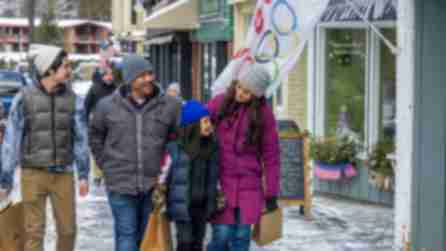 A smiling family of four walks together on a snowy sidewalk, holding shopping bags. They wear winter clothing. An Olympic flag waves nearby.