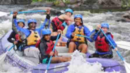 A group of people in red and orange life jackets float down lively white water rapids in a purple inflatable raft