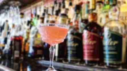 A light pink cocktail on a bar with liquor bottles on shelves