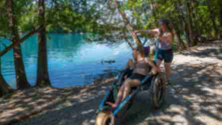 A woman pushes her daughter in a wheelchair along a lakeside path at Green Lakes State Park. The vibrant blue water, surrounded by trees, adds a serene and joyful tone.