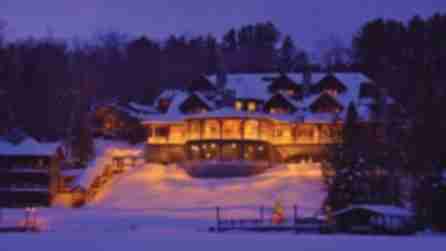 The warmly lit Lake Placid Lodge sits in a snowy landscape at dusk. Soft golden lights create a cozy glow, contrasting with the deep blue sky and snow.