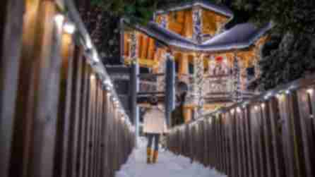 A person in a hat, white jacket, jeans, and tan boots walks down a snow-covered path toward a wooden treehouse covered in white lights