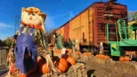 A scarecrow standing amid hay bales and pumpkins with a train in the background