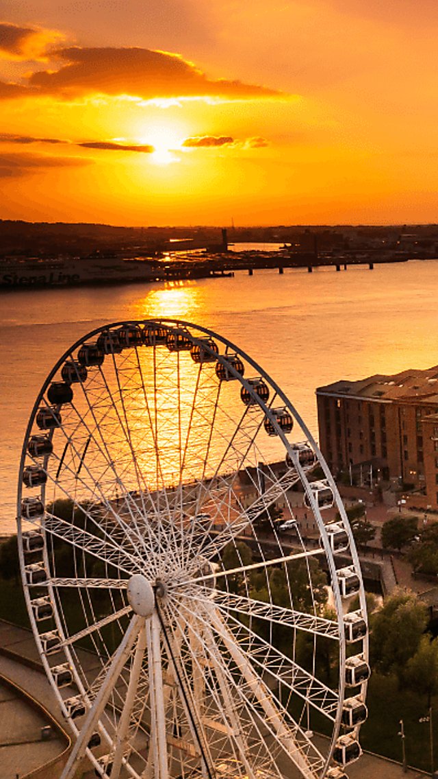 A drone view above the Liverpool Wheel ferris wheel, the Royal Albert Dock buildings and acoss the river as the sun is setting behind the Wirral.