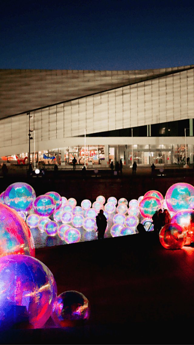 Several bubbles lit up in the river with people looking at them. It's a dark sky and the bubbles are illuminating the area.
