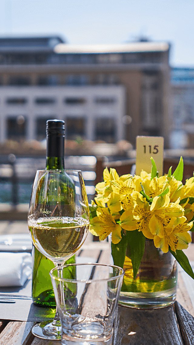 A table alongside one of the Liverpool docks, outside the Malmaison Hotel. It is a wooden outdoor table with a bunch of yellow flowers on the middle of the table, a bottle of white wine and table settings.