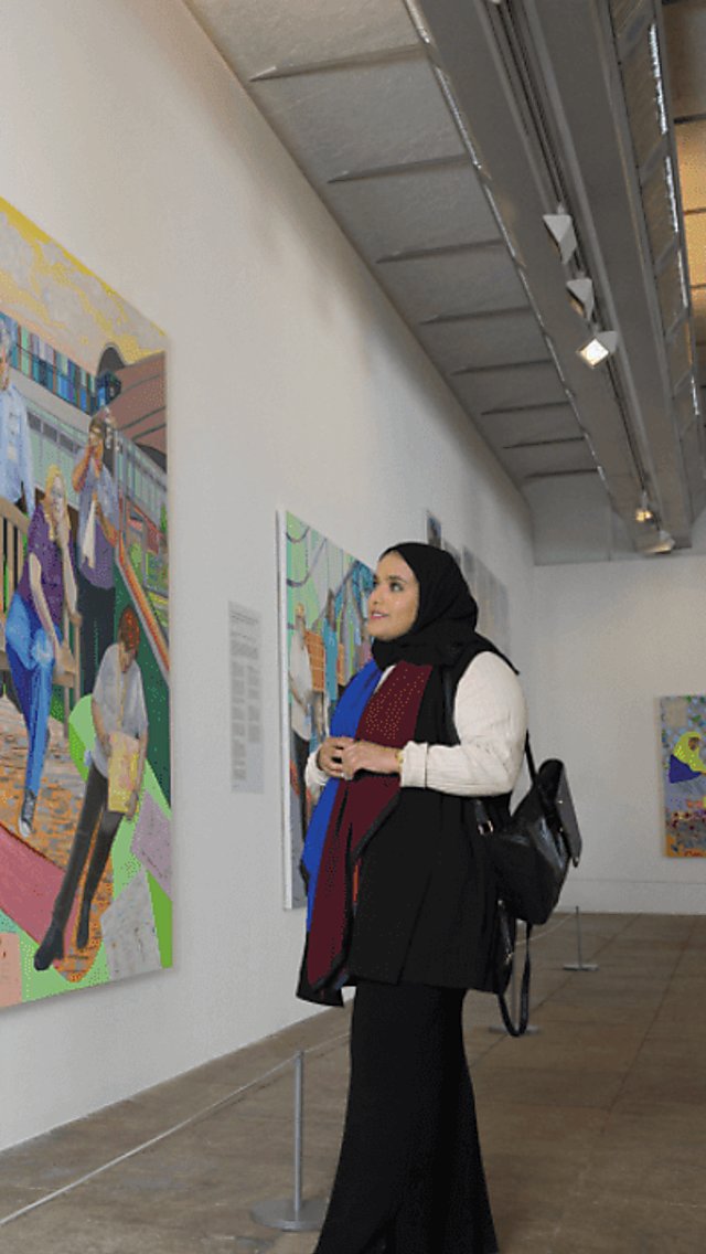 A lady wearing long black clothes looks at a large colourful and lifelike painting in a gallery. The gallery has white walls and grey metal supports.
