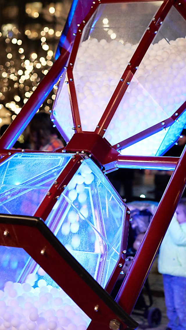 A giant hourglass filled with balls and lit up being flipped over by people engaging with the artwork