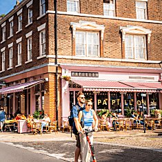 Liverpool Hope Street. The Metropolitan Cathedral is at the top of the street. To the right is a pink cafe in a Georgian Building. It is a sunny day and two people on an electric scooter.