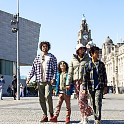 A family of four, two adults and two children walking across Liverpool Pier Head with the Royal Liver Building in the background.
