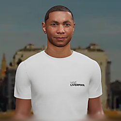 A centered, front-facing portrait of a person wearing a plain white T-shirt with the words “Visit Liverpool” printed in small black text on the upper left chest. The background is softly blurred, showing indistinct buildings and architectural shapes, giving a city setting. The person is framed from the chest up, with even lighting and a neutral backdrop that keeps focus on the face and the shirt’s text.
