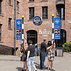 Outside of The Beatles Story with people stood by the entrance. There are blue banners either side of the staircase leading down into the attraction.