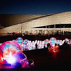 Several bubbles lit up in the river with people looking at them. It's a dark sky and the bubbles are illuminating the area.