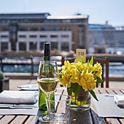 A table alongside one of the Liverpool docks, outside the Malmaison Hotel. It is a wooden outdoor table with a bunch of yellow flowers on the middle of the table, a bottle of white wine and table settings.