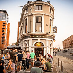 A summer evening outside the Baltic Fleet Pub. The narrow stand-alone pub building is centre of the shot, with the dock warehouse buildings to the right and modern apartments to the left. People are dressed in summer clothes sitting at wooden picnic benches.  i