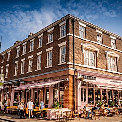 A corner shot of a Georgian Town House style building with sash windows. The ground level of the building has been turned into a cafe, painted a complimentary light pink with awnings and outdoor seating. The sky is blue and it looks like a warm summer day.