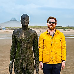 A life-size cast-iron statue on Crosby Beach in a standing position. There is a person standing next to the statue, smiling and wearing a yellow raincoat and sunglasses. They are mimicking the stance of the statue.