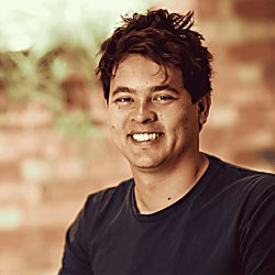 A headshot of Matt Pierri looking at a camera wearing a black t-shirt. They have brown hair and the background is a brick wall.