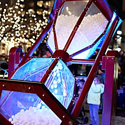 A giant hourglass filled with balls and lit up being flipped over by people engaging with the artwork