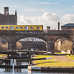 A yellow train pictured along a brick railway bridge running above a canal lock system.