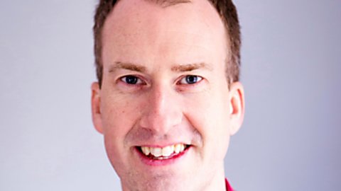 A man wearing a red shirt and blue, tartan suit jacket smiles at the camera.