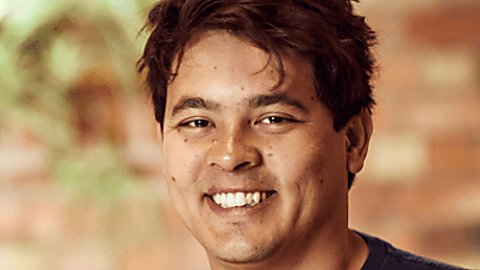 An image of a man smiling brightly and showing his teeth, his hair is black and he is wearing a navy top.