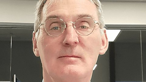 A man looking at the camera wearing a shirt and navy blue tie. He has a lanyard round his neck and is wearing glasses.