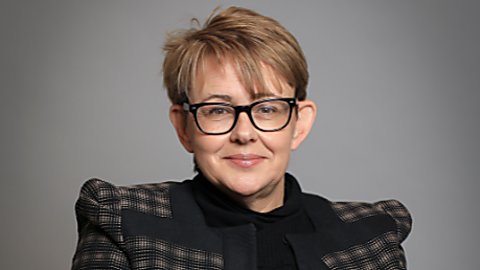 A lady wearing a black outfit including a jacket with prominent shoulder pads, smiles at the camera. She has short brown hair with highlights and black, thick framed glasses. She is sitting in a wheelchair.