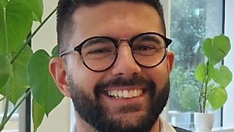 A man with well groomed, dark hair smiles at the camera form behind, black, round lense glasses. He has a short beard and is wearing a white shirt, with a navy blue jumper over.
