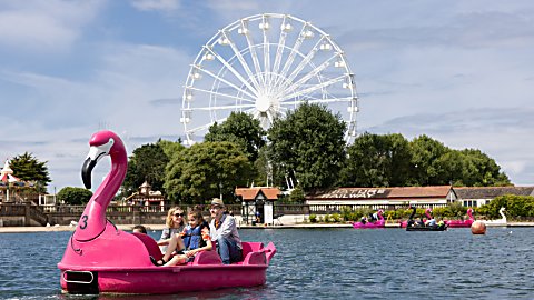 A family of two adults and two children on a pink swan boat on a marine lake in southport. There is a large ferris wheel in the background.