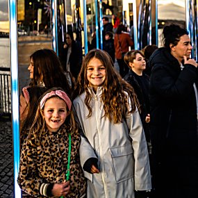 Two young girls in front of mirrored monoliths on Liverpool Pier Head. It is dusk and the Royal Liver Building can be seen in the background.