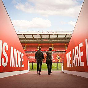 Two people are walking out of the players tunnel at Liverpool's stadium, Anfield. Either side of them are walls in red which have the words 'We are Liverpool. This means more'. In front of the two figures you can see the stand on the other side of the pitch and blue sky with few clouds.